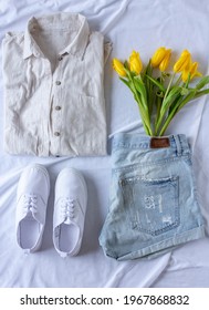 Women Clothing Flat Lay With Blue Jeans Shorts, Beige Shirt, White T Shirt, White Sneakers And Yellow Tulips, Fashion Blogger Content, Denim And Linen Texture