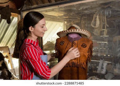 Women craftsman making leather by hand, using holding shirt made brown leather that is worn on mannequin showing shirt, there are hat made from animal skins, handmade by artisans every step way. - Powered by Shutterstock