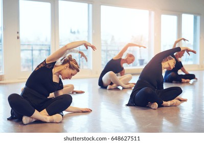 Women dancing ballet while sitting and stretching with hands up on the floor of class.