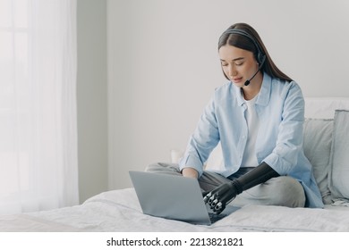 Women With Disability Wearing Headphones, Learning Online At Laptop At Home. Young Woman In Headset Typing Using Bionic Prosthetic Arm, Sitting On Bed. Lifestyle Of People With Disabilities.