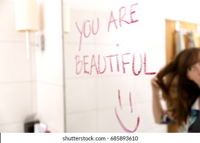 Women encouraging herself by writing words you are beautiful on mirror with lipstick
