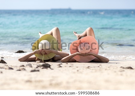 women enjoy the sea by laying down on sand of beach wearing millinery hat, looking to the swell approach to the legs