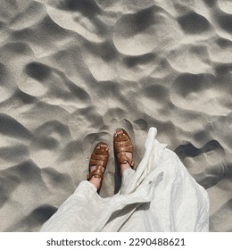 Women feet on desert beach sand. Aesthetic summer vacation background. Woman in white dress and leather sandals on the dune sand. Top view