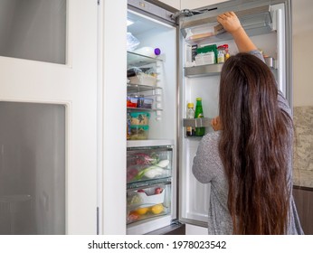 Women Grabbing Food From The Shelf Fridge. Refrigerator With Leftovers Container, Vegetables And Milk. Female With Long Hair Preparing A Meal And Searching For Ingredients. Midnight Snack.