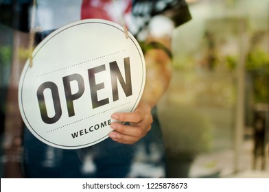 Women Hand Flip The Sign To Open The Shop. Open Sign Hanging On The Glass Door.