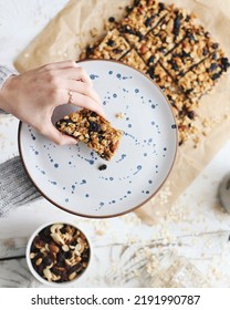 Women Hand Holding Granola Bar On A Plate With Blue Sparkles Over A Table With The Rest Of The Homemade Granola 