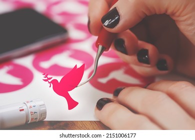 women hands with painted nails hold freshly cut peace dove decal that sticks to tip of curved weeding tool. pink adhesive film. wooden worktop. adjustable plotting blade in foreground. selective focus