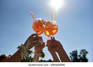 Women hands toasting with aperol spritz cocktails on summer party on blue sky background, copy space. Event celebration concept. Traditioanal italian aperitif. - Shutterstock ID 2083420096