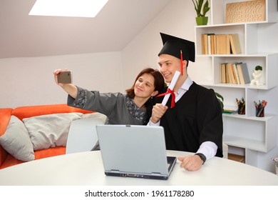 A Women And Her Son In Graduation Gown And Cap Making Selfie Photo With Mobile Phone At Home, During The Broadcast Of The Ceremony. Class Of 2021. Covid-19 New Normal Concept.