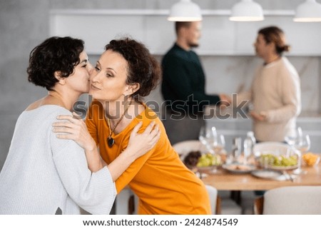 Women hug each other and kiss on the cheek while greeting each other before a festive dinner. Men shaking hands in the background
