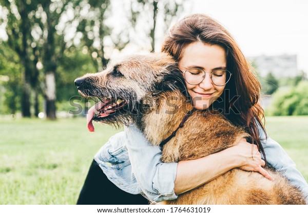 Women hugging\
dog in the summer park. Cheerful lady with long dark hair in blue\
jacket hugs and strokes friendly old dog sitting on lush green\
meadow of public garden on nice day.\
