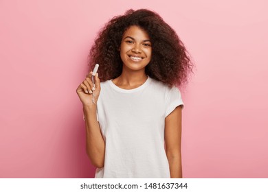 Women Hygiene Protection Concept. Cheerful Dark Skinned Female Holds Cotton Tampon, Satisfied With Good Protection During Critical Days, Wears Casual White Mockup T Shirt, Isolated On Pink Wall