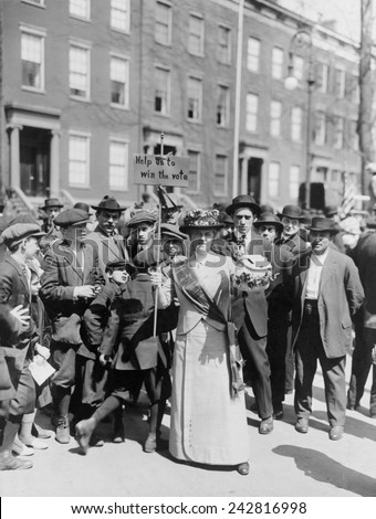 Women identified as Mrs. Suffern, is surrounded by a crowd of men and boys, while she holds a home-made banner in women suffragist parade 'Help us to win the vote.' 1914.