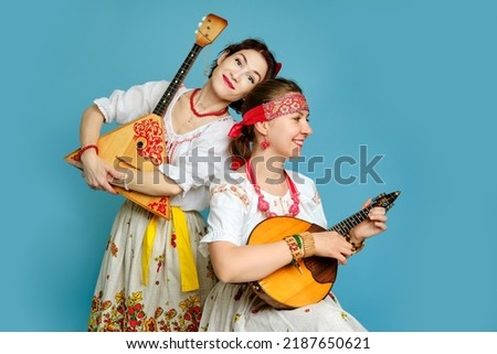 Women musicians in Russian folk dresses with musical instruments on a blue studio background. Happy artists from Russia in white national clothes with stringed musical instruments
