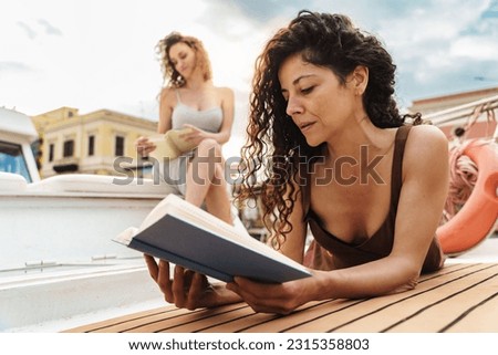 Women Reading on Boat Deck - A Caucasian woman with curly hair engrossed in reading, while her Brazilian friend relaxes and reads. Enjoying the sun and the boat's ambiance.