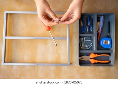 Women Repairing An Old Metal Object. Red And Yellow Screwdriver, Orange Pliers, Screws And Blue Tape Measure In Toolbox. Yellow Wooden Workshop Bench. Top View Photo. Home Hobby In Lockdown.