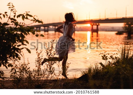 Women running at sunset. Lifestyle full-length portrait of a beautiful young woman in a long white dress running along the riverbank at sunset. 