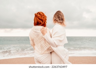 Women sea walk friendship spring. Two girlfriends, redhead and blonde, middle-aged walk along the sandy beach of the sea, dressed in white clothes. Against the backdrop of a cloudy sky and the winter - Shutterstock ID 2263763497
