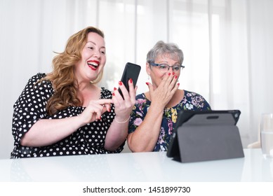 Women Sitting At A White Table Using Tablet And Smart Phone In Front Of White Curtains In The Background. Overweight And Mature Woman Looking At Screen Surprisedly And Shocked With Hand In Front Of Mo