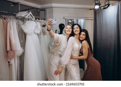 Women Taking Photographs Of A Female Friend Trying On Wedding Dress. Women In Wedding Dress Fitting Room.