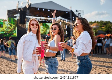 Women toast with cocktails at sunny beachside music event, enjoying performance, friendship, and leisure. Smiling attendees in summer attire with drinks, relish outdoor festival vibe near ocean. - Powered by Shutterstock