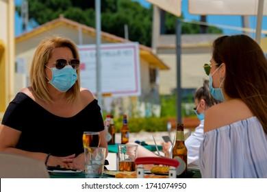 Women Using Surgical Masks And Sunglasses Sitting At An Outdoor Bar Terrace With Beers And Snacks On An Out Of Focus Background. New Lifestyle Concept.