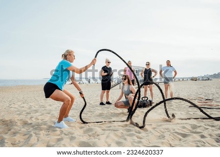 Women of various ages doing fitness workouts in class exercise with coach on the beach. Middle aged lady working with battle ropes. Intense functional circuit training. Sport for health and wellbeing