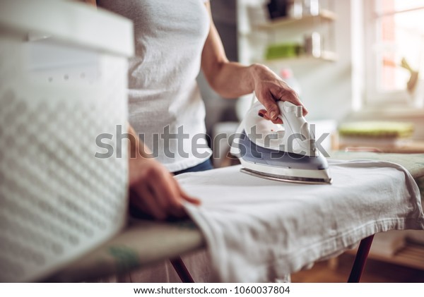 Women wearing white shirt ironing clothes on\
ironing board in laundry room at\
home
