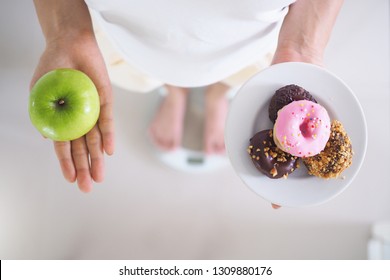 Women Are Weighing The Body On The Scale. Holding Donut Dish And Apple. The Difficulty Of Having To Refrain From Foods That Contain Trans Fat, Lose Weight, Do Not Eat Flour. Eating Fruit For Health 