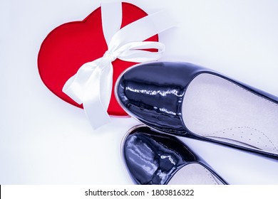 Women's black patent leather shoes, one of which stepped on a red silk box in the shape of a heart with a white ribbon 