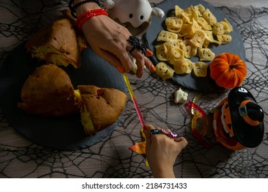 Women's And Child's Hands With Spider Rings And Braceletes Are Pulling The Jelly Gummy Worm. Halloween Trick Or Treat Composition With Ghost Chips, Pumpkin Cake And Spiderweb Tablecloth. Top View.