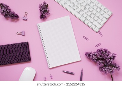 Women's desktop close-up, keyboard, notebooks with pen, lilac flowers. Minimal cropped flat composition on a pink background. Copy space