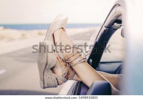 Women\'s feet out of the car window with a road\
and the sea in background. Crossed legs of young girl with trendy\
shoes. Female relaxing during summer adventure. Freedom, travel,\
female\'s fun concept.