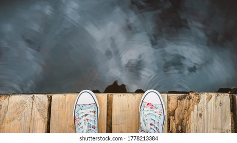 Women's feet stand on the edge of a wooden dock. A girl in sneakers is resting on the lake