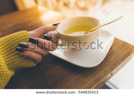 Women's hand with a manicure hold a cup of tea near winter window, on wooden table
