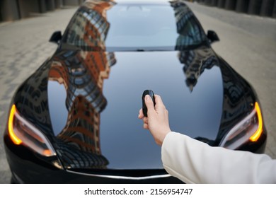 Women's Hand Pressing On Remote Control Automobile Alarm Systems. Female Arm Holding Car Key On The Background Of Luxure Modern Black Auto.