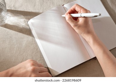 Women's hand writing in a blank notebook sheet with sunlight shadows, home office lifestyle workspace, mock up with copy space