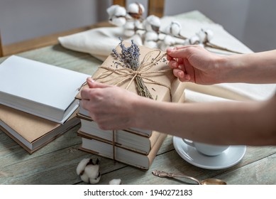 Women's hands is carefully decorating a stack of books with craft paper covers with rope and lavender flowers. Concept of packaging books for a gift.