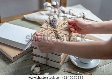 Women's hands is carefully decorating a stack of books with craft paper covers with rope and lavender flowers. Concept of packaging books for a gift.