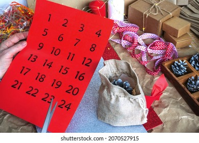 Women's hands cut the numbers for the advent calendar, on the table there are gift packages and a ready-made gift in a canvas bag, cones, ribbons. Preparing for Christmas. DIY
