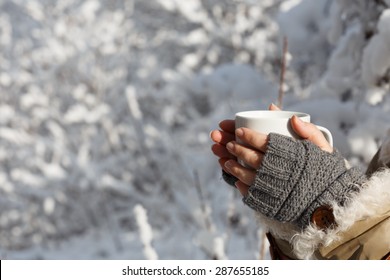 Women's hands in gray fingerless knitted mittens and coat with white fur holding white cup of tea at the background of snowy forest. Close up.