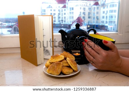 women's hands hold a large black cup of tea with a kettle next to it, cookies in a plate and an open book on the background of a window overlooking the city.