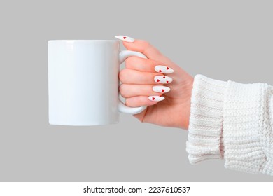 Women's hands hold a mockup of a white empty mug on a gray background, a cup for your design and logo close-up. White manicure nails with red hearts for Valentine's Day.