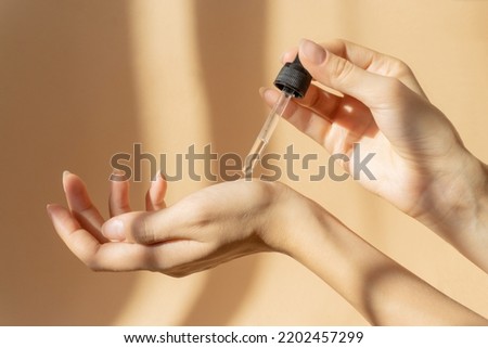 Women's hands hold pipette with cosmetic serum on light brown background, in rays of sunlight. Close-up, copy space. Concept of body care