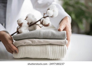 Women's hands hold a stack of neat, clean, cotton clothes in neutral shades. Branch with cotton inflorescences. Organic cotton concept.