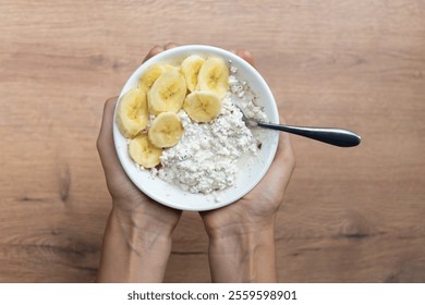 Women's hands hold a white bowl with cottage cheese, banana and fiber on a wooden table, ideal for healthy eating.