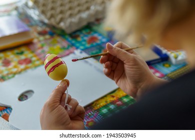 Women's hands holding an Easter egg  A brush nearly paints the yellow egg and red paint  In the blurred background you can see other painting utensils  Look over Shoulder  
