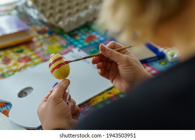Women's hands holding an Easter egg  A brush paints the yellow egg and red paint  In the blurred background you can see other painting utensils  Look over shoulder  