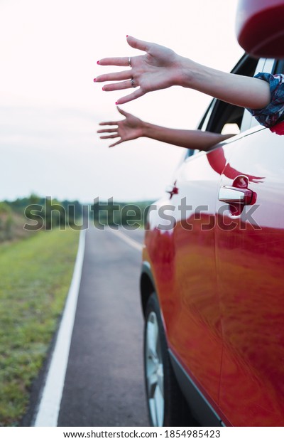 Women\'s hands outside the car as if caressing the\
wind - The girls lean out of a car window against the backdrop of\
the long road.