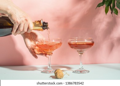 Women's Hands Pour Champagne Or Wine Into Transparent Elegant Glasses. Pink Pastel Background Bright Light. Copy Space.